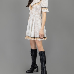 collector of vision model is wearing a size small white lace baby doll dress showing the side with western gold and black trim, mock neckline, short puff sleeves, fit and flare ruffle hemline, styled with black tall boots