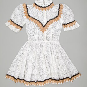 collector of vision model is wearing a size small white lace baby doll dress with western gold and black trim, short puff sleeves, fit and flare ruffle hemline