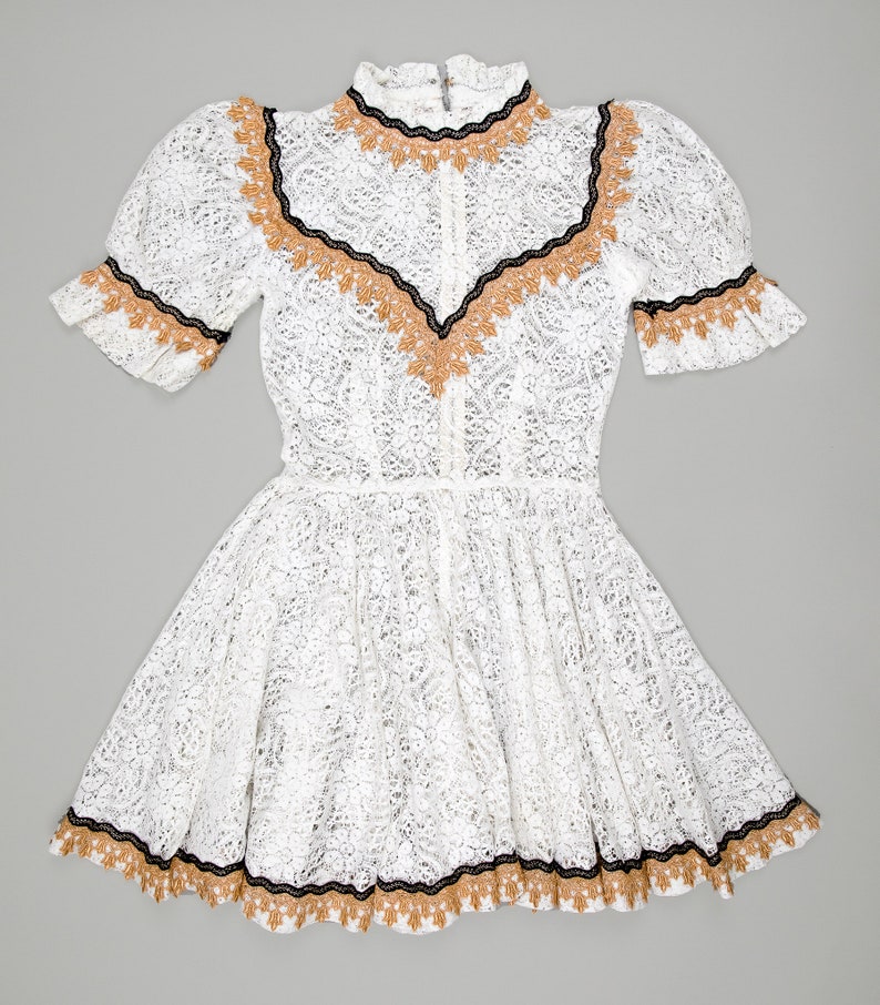 collector of vision model is wearing a size small white lace baby doll dress with western gold and black trim, short puff sleeves, fit and flare ruffle hemline