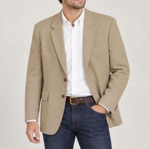 May include: A tan blazer with notched lapels and two front buttons, worn over a crisp white button-down shirt. The outfit includes dark blue denim jeans secured with a brown leather belt featuring a gold-toned rectangular buckle.