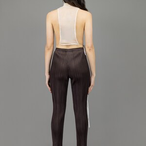collector of vision model is wearing a size small victorian lace collar vest showing off the back with mesh, adjustable side tie closure, styled with plum issey miyake pleated pants, and nude pumps