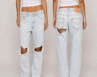 May include: Light-wash blue denim jeans featuring a high-waisted fit, a button fly, and distressed detailing with large rips at both knees. The back view displays two rear pockets, a tan leather-style brand patch, and a large tear under the right pocket.