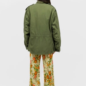 collector of vision model is wearing a size medium 70s wide floral pants showing off the back, styled with green military cargo jacket, white pointed kitten heels, and crystal dangling earrings