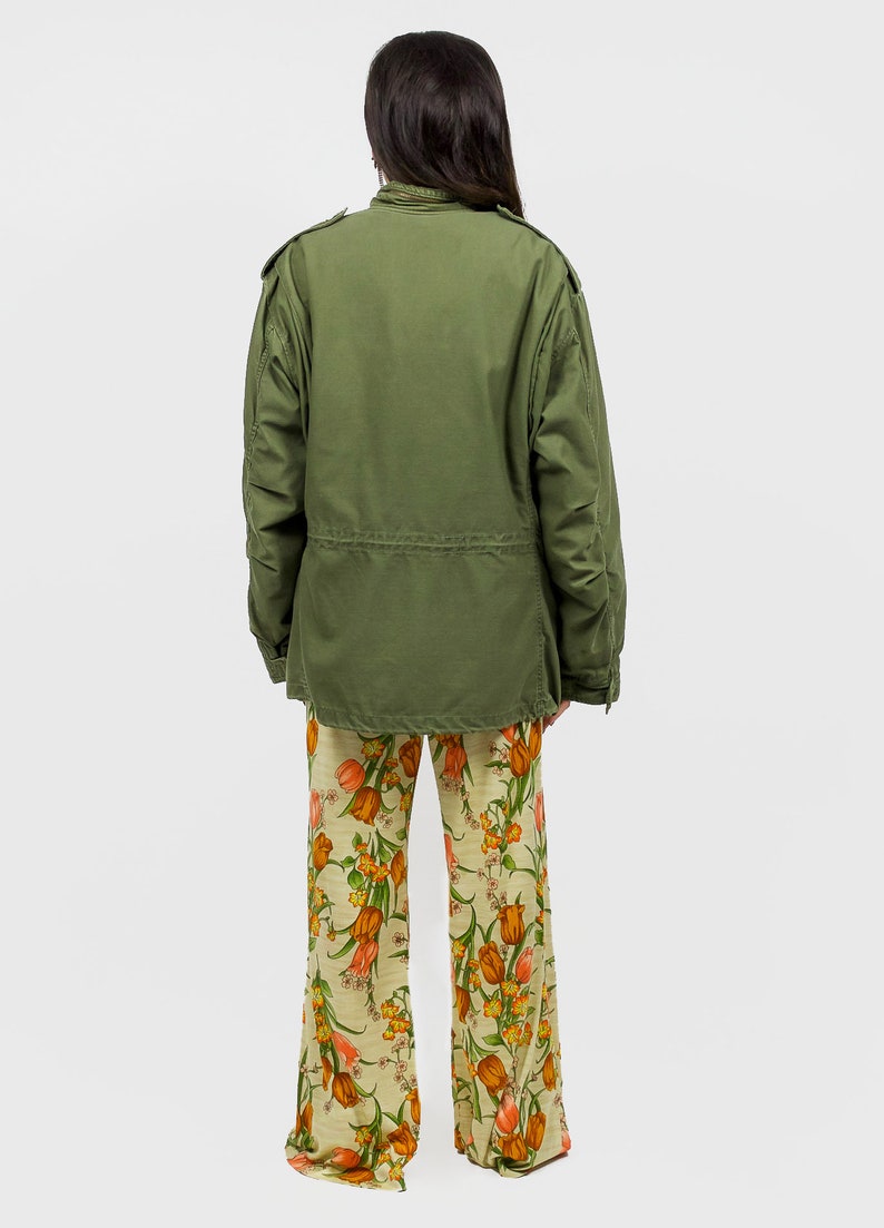 collector of vision model is wearing a size medium 70s wide floral pants showing off the back, styled with green military cargo jacket, white pointed kitten heels, and crystal dangling earrings