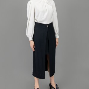 collector of vision model is wearing a size medium black short long midi skirt showing off the side with silver decorative button, short and long length, styled with a formal white shirt, plum buckle pumps, and crystal dangling earrings