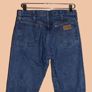 K&ouml;nnte beinhalten: Eine dunkelblaue Jeanshose mit einem braunen Leder-Patch auf der Ges&auml;&szlig;tasche, auf dem "Wrangler" steht.