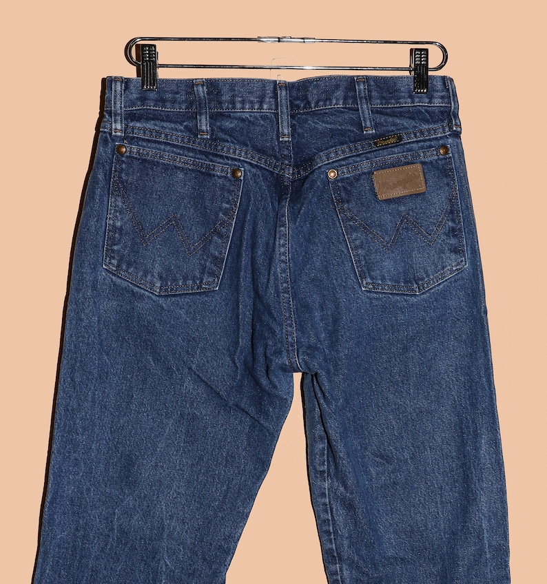 K&ouml;nnte beinhalten: Eine dunkelblaue Jeanshose mit einem braunen Leder-Patch auf der Ges&auml;&szlig;tasche, auf dem "Wrangler" steht.