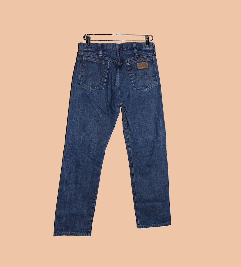 K&ouml;nnte beinhalten: Eine dunkelblaue Jeanshose mit einem braunen Leder-Patch auf der Ges&auml;&szlig;tasche. Die Jeans h&auml;ngt an einem Kleiderb&uuml;gel.