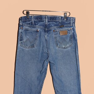 Pu&ograve; includere: Un paio di jeans blu in denim con una toppa in pelle marrone sulla tasca posteriore che dice "Wrangler".