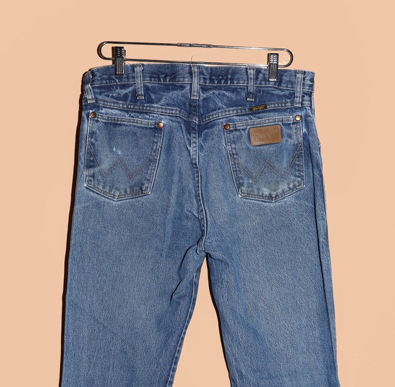 Pu&ograve; includere: Un paio di jeans blu in denim con una toppa in pelle marrone sulla tasca posteriore che dice "Wrangler".