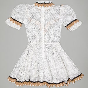 collector of vision model is wearing a size small white lace baby doll dress showing off the back side with western gold and black trim, short puff sleeves, fit and flare ruffle hemline, back zipper closure