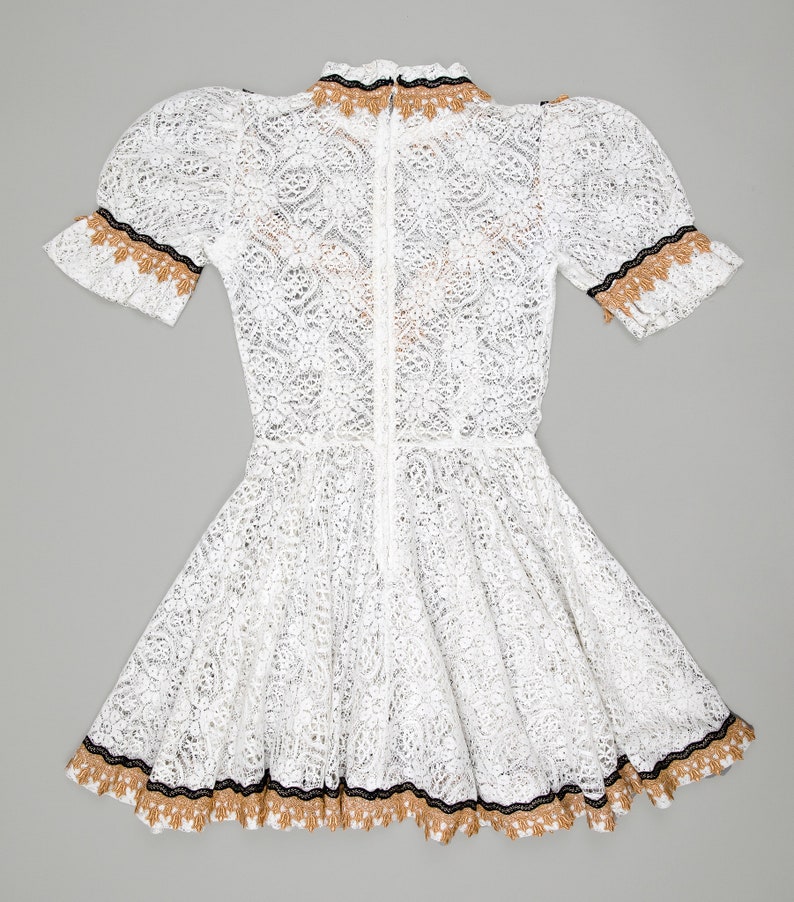 collector of vision model is wearing a size small white lace baby doll dress showing off the back side with western gold and black trim, short puff sleeves, fit and flare ruffle hemline, back zipper closure