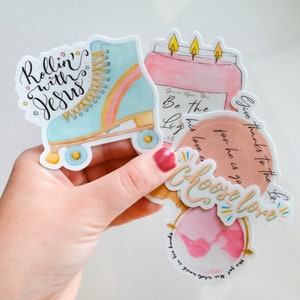 May include: Four colorful stickers with inspirational messages. The stickers feature a roller skate, a candle, a globe, and a heart. The roller skate is blue and pink with a rainbow stripe. The candle is pink with three yellow flames. The globe is pink with a white outline. The heart is pink with a white outline. The text on the stickers reads "Rollin' with Jesus", "Be the Light", "Give thanks to the Lord", and "Choose Love".