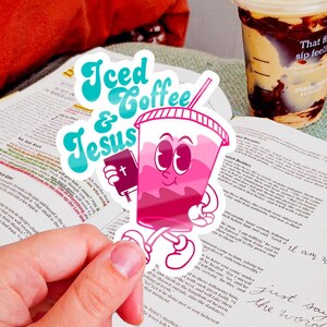 May include: A pink sticker with the words "Iced & Jesus" and a cartoon iced coffee cup holding a bible. A cup of coffee with chocolate swirls and the text "That first sip feeling" is visible. An open book is in the background.