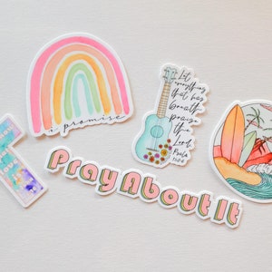 Pode incluir: Um conjunto de cinco adesivos coloridos com um arco-&iacute;ris, uma cruz, um ukulele, uma prancha de surf e as palavras "Ore por isso". Os adesivos est&atilde;o todos em um fundo branco.