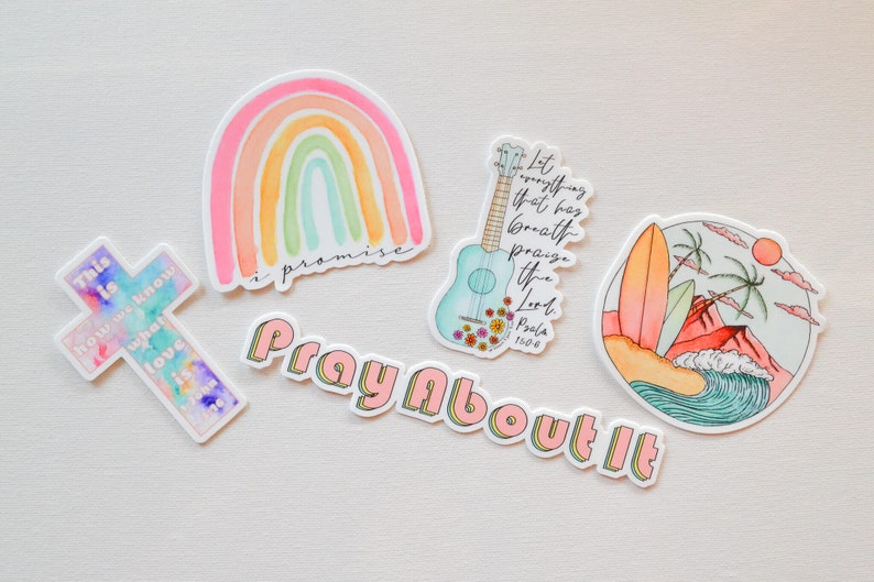Pode incluir: Um conjunto de cinco adesivos coloridos com um arco-&iacute;ris, uma cruz, um ukulele, uma prancha de surf e as palavras "Ore por isso". Os adesivos est&atilde;o todos em um fundo branco.