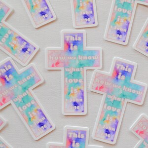 Pode incluir: Um conjunto de adesivos em forma de cruz coloridos com o texto "This is how we know what love is 1 John 3:16". As cruzes s&atilde;o pintadas em estilo aquarela com tons de rosa, azul e verde.