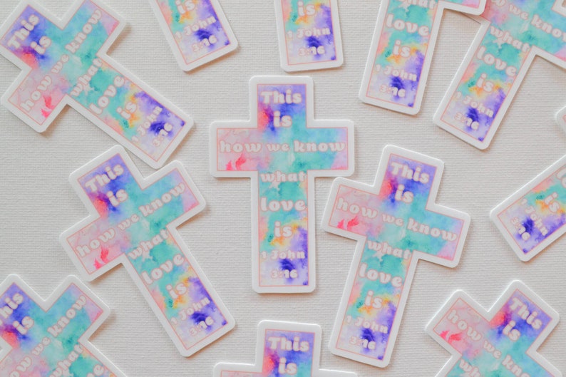Pode incluir: Um conjunto de adesivos em forma de cruz coloridos com o texto "This is how we know what love is 1 John 3:16". As cruzes s&atilde;o pintadas em estilo aquarela com tons de rosa, azul e verde.
