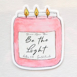 May include: A pink candle jar with a white label that reads "Grace Upon You Be the Light Matthew 5:16 Scented Candle". The candle has three yellow flames.