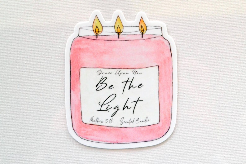 May include: A pink candle jar with a white label that reads "Grace Upon You Be the Light Matthew 5:16 Scented Candle". The candle has three yellow flames.