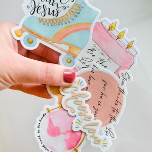 May include: A set of five watercolor stickers with inspirational quotes. The stickers feature a roller skate, a candle, a globe, and two other designs. The quotes on the stickers are "Rollin' with Jesus", "Give thanks to the Lord for he is good", "Be the light", "Grace upon you", and "Choose love".
