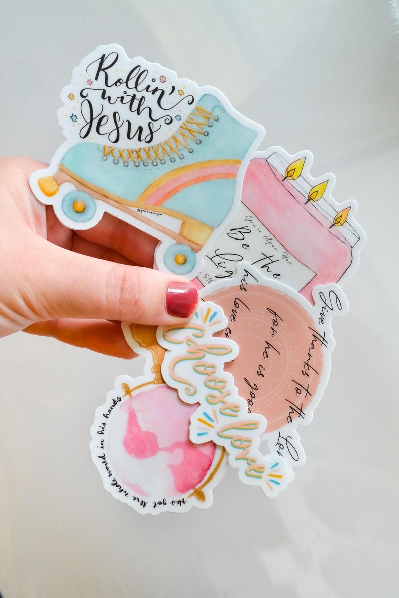 May include: A set of five watercolor stickers with inspirational quotes. The stickers feature a roller skate, a candle, a globe, and two other designs. The quotes on the stickers are "Rollin' with Jesus", "Give thanks to the Lord for he is good", "Be the light", "Grace upon you", and "Choose love".
