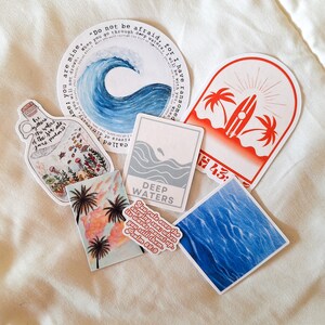 May include: Assortment of stickers featuring ocean and beach themes. Stickers include a blue wave with the text "Do not be afraid, for I have ransomed you. When you go through deep waters, I will be with you, you will not drown. You are mine."  Another sticker features a palm tree and surfboard with the text "H43". A third sticker features a blue ocean wave with the text "DEEP WATERS" and the scripture "You rule over the surging sea; when its waves mount up, you still them. Psalm 89:9".