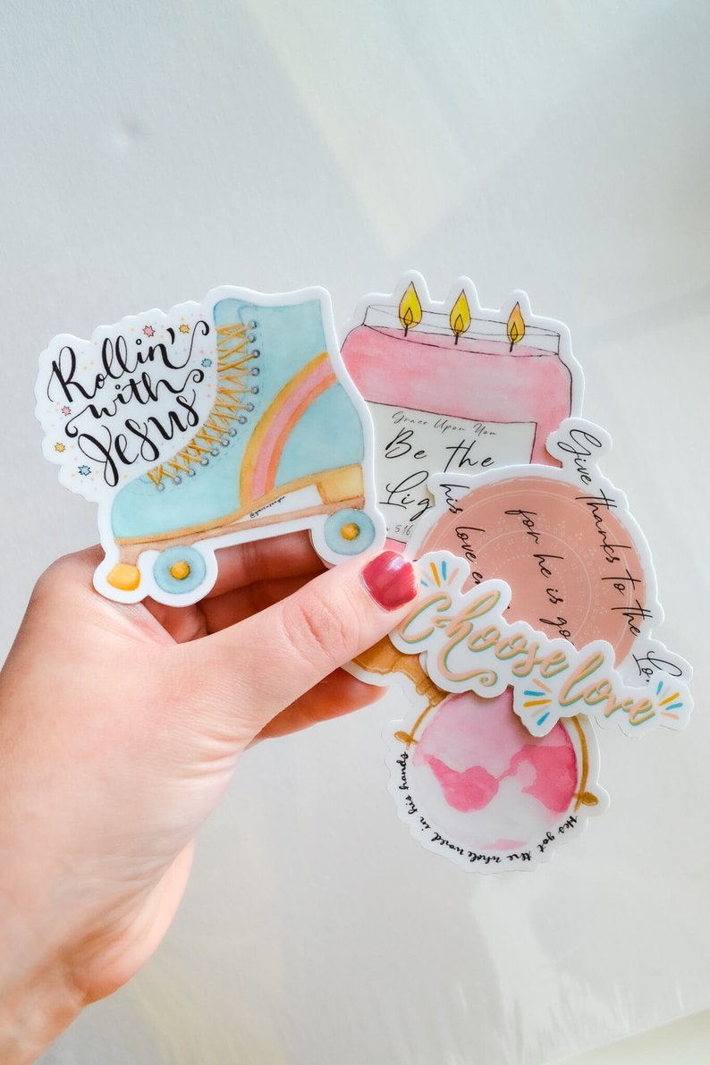 May include: Four colorful stickers with inspirational messages. The stickers feature a roller skate, a candle, a globe, and a heart. The roller skate is blue and pink with a rainbow stripe. The candle is pink with three yellow flames. The globe is pink with a white outline. The heart is pink with a white outline. The text on the stickers reads "Rollin' with Jesus", "Be the Light", "Give thanks to the Lord", and "Choose Love".