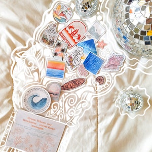May include: A collection of colorful stickers with various designs, including a seashell, a surfboard, a bottle, and a palm tree. The stickers are arranged on a white surface with a white outline around them. There are two silver disco balls in the background.