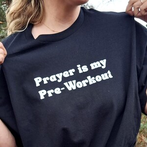 May include: Black t-shirt with the white text "Prayer is my Pre-Workout". The shirt is made of a soft material and has a crew neck. The image shows the shirt being held up by a person.