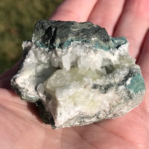 May include: A close-up of a geode with a white and green crystal interior. The geode is surrounded by a dark gray, rough exterior.