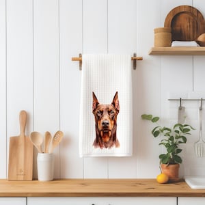May include: A white waffle-textured kitchen towel featuring a centered, detailed watercolor-style illustration of a brown and black Doberman Pinscher dog head facing forward with alert, pointed ears and a focused expression.
