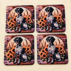 May include: A set of four square coasters featuring a repeating illustration of a black and white spotted dog sitting in front of a cluster of yellow sunflowers and wheat stalks, all set against a circular frame and a dark brown wooden plank background.