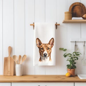 May include: A white waffle-textured kitchen towel featuring a detailed watercolor illustration of a brown and white Basenji dog head. The dog has pointed ears, amber eyes, and a white muzzle, centered on the fabric hanging from a wooden rod.