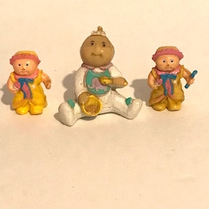 May include: Three plastic Cabbage Patch Kids dolls. One doll is wearing a white onesie with a blue bib and a yellow hat. Another doll is wearing a yellow dress with a pink bow and a yellow hat. The third doll is wearing a yellow dress with a pink bow and a pink hat.