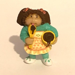 May include: A Cabbage Patch Kid doll figurine wearing a teal blue sweater and a yellow and pink apron. The doll is holding a yellow spoon and a brown bowl with chocolate inside.
