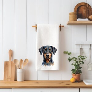 May include: A white waffle-textured kitchen towel featuring a centered watercolor illustration of a Doberman Pinscher dog. The dog has a black coat with tan markings on its face, chest, and muzzle, depicted with detailed brushstrokes.