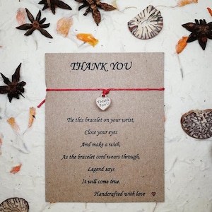 Könnte beinhalten: Eine Dankeskarte mit einem roten Schnurarmband und einem Herzanhänger. Die Karte trägt die Worte "THANK YOU" und eine Botschaft über das Wünschen. Der Hintergrund umfasst Sternanis und getrocknete Blütenblätter.