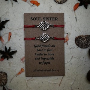 May include: Two red string bracelets with silver knot charms. The bracelets are packaged on a brown card with the text "SOUL SISTER" and a quote about good friends. The card is handcrafted with love.