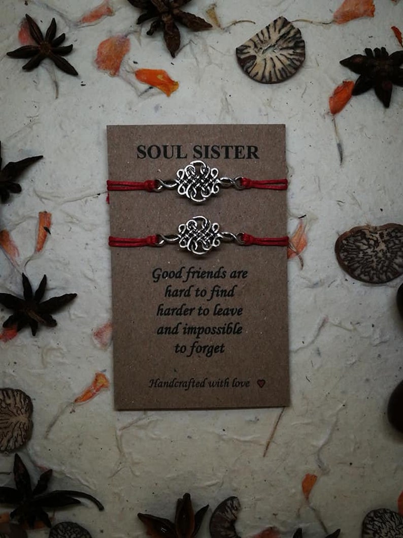 May include: Two red string bracelets with silver knot charms. The bracelets are packaged on a brown card with the text "SOUL SISTER" and a quote about good friends. The card is handcrafted with love.