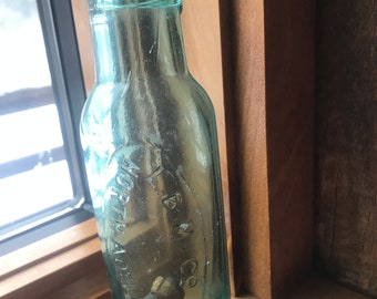 May include: A vintage green glass bottle with a cork top. The bottle has a label that reads "T.B.M. Co. Appleton, Wis."