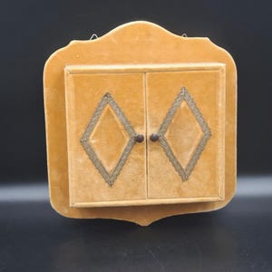 May include: A wall-mounted cabinet with a soft, mustard-yellow velvet finish. The front features two doors, each decorated with a central diamond-shaped inlay outlined in metallic gold trim and a small, dark circular knob for opening the doors.