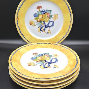 May include: A set of yellow and white ceramic plates. Each plate features a yellow border with a blue ribbon design. The center of each plate has a floral and fruit bouquet with a blue ribbon.