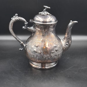 Puede incluir: Una tetera de metal plateado con un patrón floral y de volutas en relieve en su cuerpo redondeado. Presenta un asa curva con textura detallada, un pico a juego y una tapa rematada con un pequeño remate floral esculpido en la parte superior.