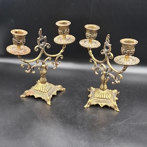 May include: A pair of two-armed brass candelabras featuring ornate scrollwork, square fluted bases, and textured candle cups. Each metal candle holder has a central stem with two curved arms extending outward to support circular drip plates.