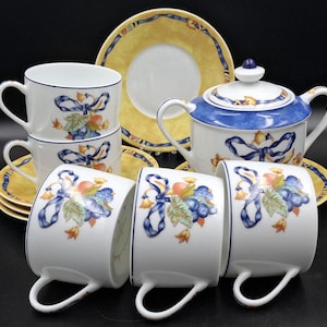 May include: A white porcelain tea set with a blue and yellow floral design. The set includes cups, saucers, a teapot, and a sugar bowl. The cups and teapot feature a blue ribbon and fruit motif. The saucers and teapot lid have a yellow border.