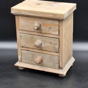 Può includere: Una piccola cassettiera rettangolare in legno con tre cassetti sovrapposti, ciascuno dotato di un pomello rotondo in legno. L'oggetto è realizzato in legno chiaro non rifinito con venature e texture visibili, poggiante su quattro piedini.