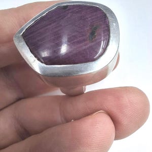 May include: A ring featuring a large, irregular, pear-shaped purple stone set in a thick, polished silver band. The stone displays a deep violet hue with visible linear striations and a small dark inclusion on the right side of its smooth surface.
