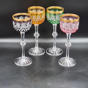 May include: Four crystal wine glasses featuring a cut glass pattern and gold rims. Each glass has a distinct coloured bowl in purple, amber, green, and pink, supported by a clear stem with a spherical node and a starburst-cut circular base.