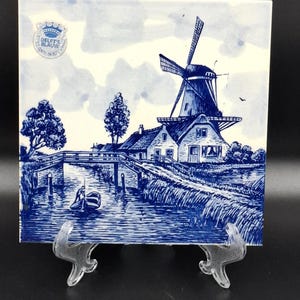 Può includere: Piastrella quadrata in ceramica con un'illustrazione blu e bianca di un mulino a vento olandese, case e un ponte su un canale. La piastrella ha un timbro Delft's Blauw. L'opera raffigura un sereno paesaggio rurale con alberi e una piccola barca.